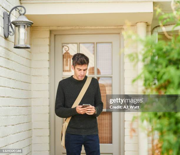 young man standing outside of his front door and texting on his phone - voordeur stockfoto's en -beelden