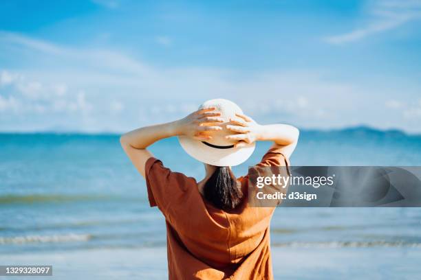 rear view of carefree young asian woman with straw hat relaxing on the beach against blue sky on a sunny summer day - orange hat stock pictures, royalty-free photos & images