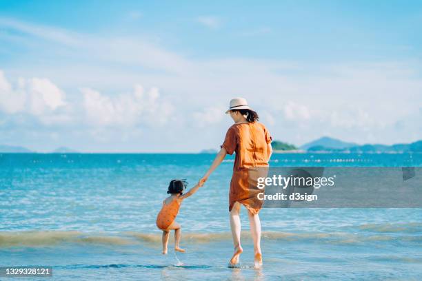 rear view of joyful young asian mother and little daughter holding hands while jumping on the beach against blue sky on a sunny summer day - family holidays hotel stockfoto's en -beelden