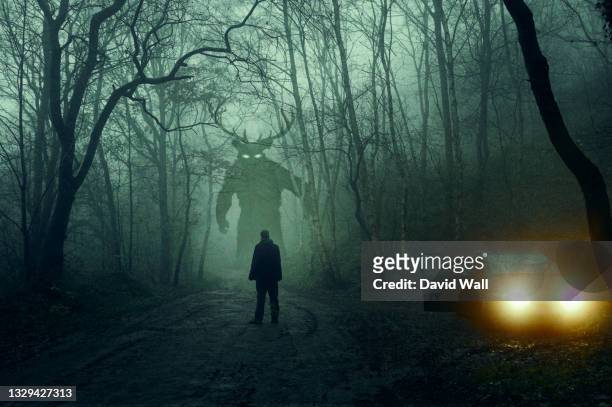 a horror concept. of a man next to a car looking at a monster with glowing eyes. in a spooky, winter forest at night with a grunge, textured edit - monstro imagens e fotografias de stock