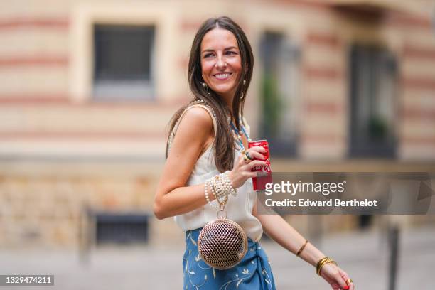 Alba Garavito Torre wears a white tank top, a beaded bracelet, a metallic sphere shaped bag, a blue floral print skirt, bejeweled rings and holds a...