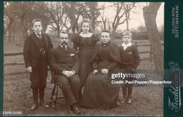 Cabinet card showing a family group comprising a man, woman and three children, the woman wears a dark, striped dress with high lace collar and gigot...