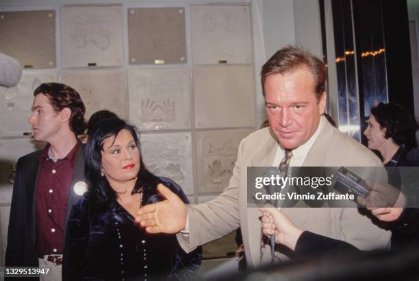 American comedian Roseanne Barr, wearing a velvet outfit with a beaded necklace, while her husband, American actor and comedian Tom Arnold gives an...