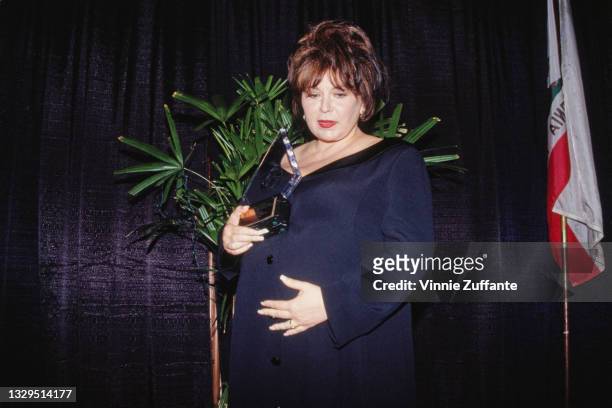 American comedian Roseanne Barr, holding an unspecified award to her chest with her right hand while cradling her 'baby bump' with her left hand,...