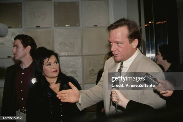 American comedian Roseanne Barr, wearing a velvet outfit with a beaded necklace, while her husband, American actor and comedian Tom Arnold gives an...