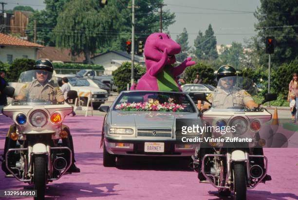 Barney, the purple Tyrannosaurus rex from children's television series 'Barney & Friends', riding in a convertible car following behind police...