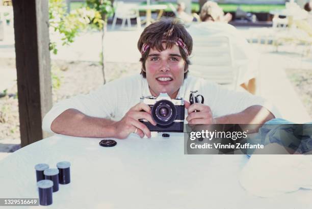 American actor Peter Barton, wearing a headband and a white shirt poses holding a Canon SLR camera, canisters for rolls of film on the table before...