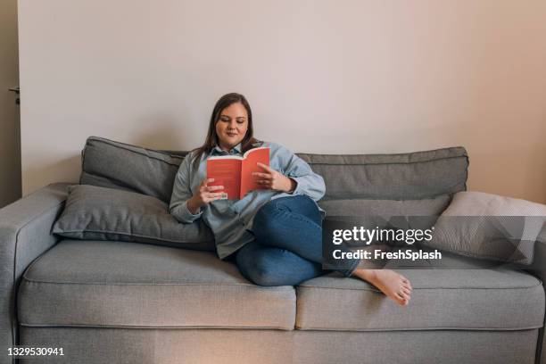 hermosa mujer de talla grande relajándose en el sofá y leyendo un libro - hourglass books fotografías e imágenes de stock