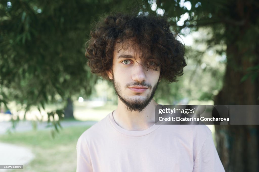 Portrait of a young man 20 years old with curly hair in a pink T-shirt on a tree background