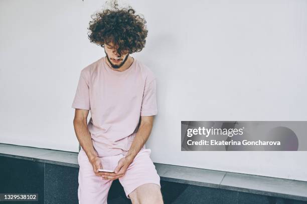 a young curly-haired man of 19 years old is sitting outside in the shade with a phone. - 20 24 years photos foto e immagini stock