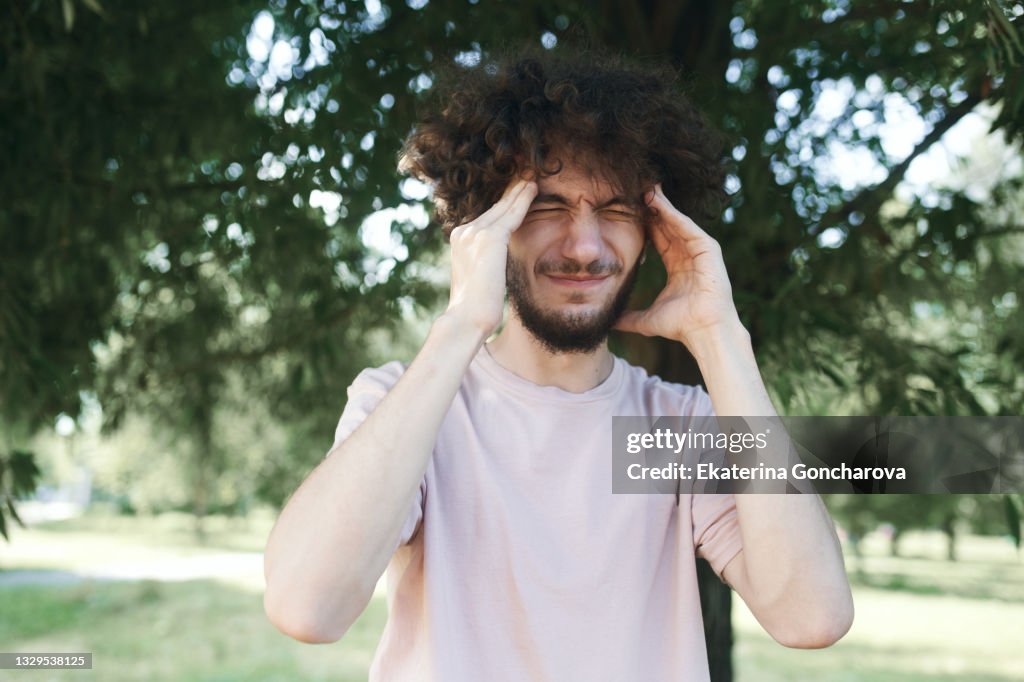 Portrait of a young man 20 years old with curly hair in a pink T shirt squeezing his head from a headache