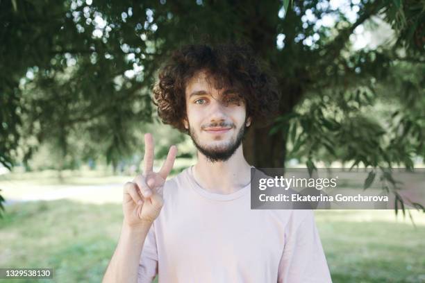 portrait of a young man with long curly hair in pink casual clothes in a city park. - victory sign stock-fotos und bilder