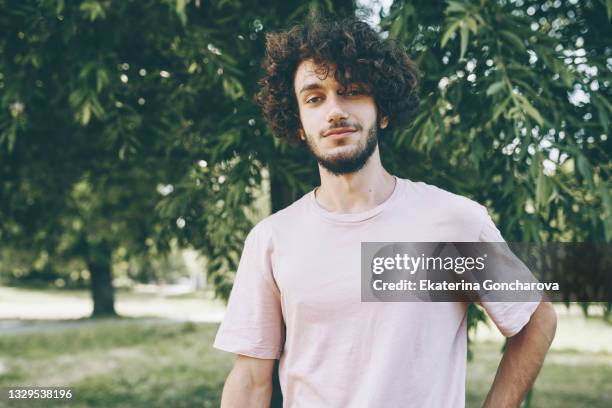 portrait of a young man with long curly hair in pink casual clothes in a city park. - curly man stock pictures, royalty-free photos & images