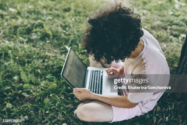 a young guy is sitting by a tree with a laptop in the summer on a green lawn - man-sitting-on-grass-using-laptop-elevated-view stock pictures, royalty-free photos & images