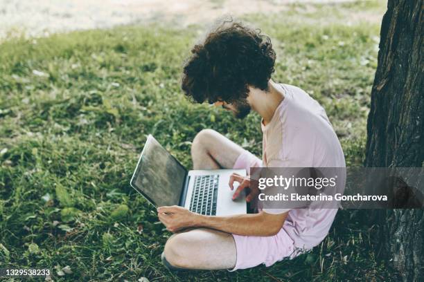 a young guy is sitting by a tree with a laptop in the summer on a green lawn - man-sitting-on-grass-using-laptop-elevated-view stock pictures, royalty-free photos & images