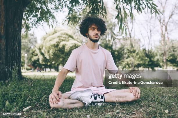 a young man with long curly hair is sitting in the lotus position and meditating in a city park. - man doing yoga in the morning photos et images de collection