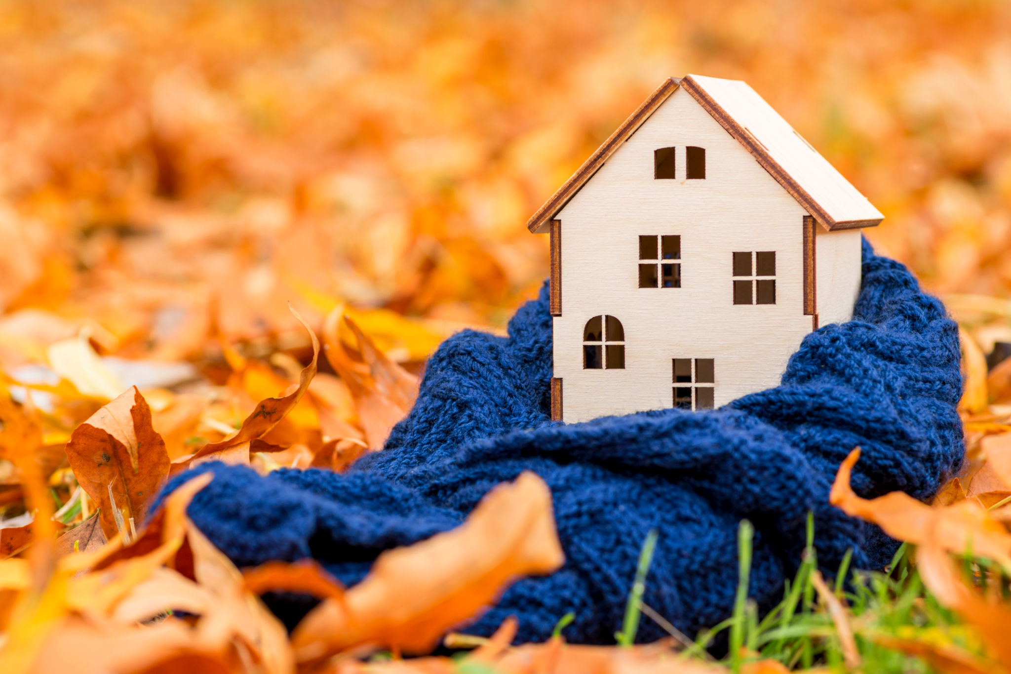 Toy wooden house is wrapped in a warm scarf on the autumn leaves background. Warm house concept. Preparing the house for the cold. Toy wooden house is wrapped in a warm scarf on the autumn leaves background. Warm house concept. Preparing the house for the cold.