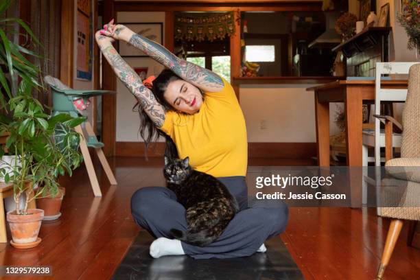 tattooed woman stretching at home with cat in lap - yoga stock-fotos und bilder