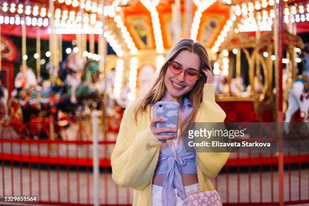 a cheerful smiling happy teenage millennial girl in casual clothes and bright pink sunglasses communicates typing using mobile phone technologies at the carousel in the amusement park at night - candid forum 個照片及圖片檔