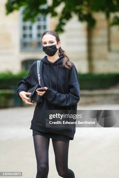 Model wears silver earrings, a black oversized hoodie sweaters, black tights, a black skirt, a black shiny leather shoulder bag, silver rings,...