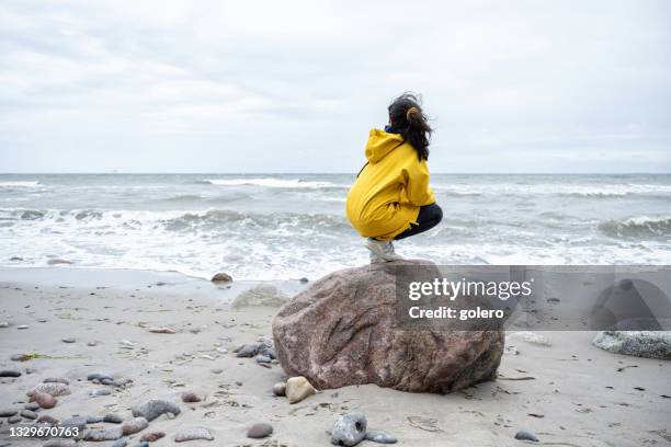 kleines mädchen in gelbem regenmantel, das auf stein kauert und das meer beobachtet - beach stone stock-fotos und bilder