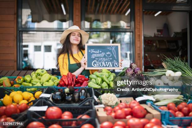 portrait of female greengrocer smiling and holding a sign in front of a grocerie market - market retail space stock pictures, royalty-free photos & images