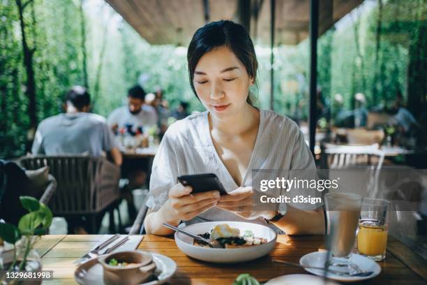 smiling young asian woman using smartphone while having brunch in an outdoor restaurant surrounded by lush foliage and beautiful sunlight. lifestyle and technology - eating yummy stock-fotos und bilder