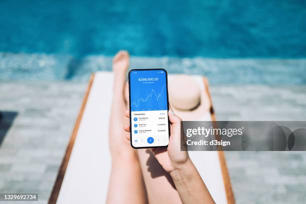 low section of young woman managing online banking with mobile app on smartphone while enjoying the sun on lounge chair by pool side. tracking and planning spending. transferring money, paying bills, checking account balance. smart banking with technology - booking hotel foto e immagini stock