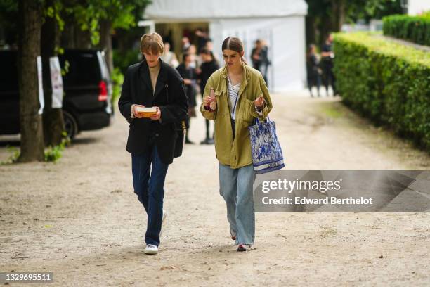 Model wears a beige wool turtleneck pullover, a black oversized blazer jacket, a black shiny leather shoulder bag, gold rings, blue denim jeans...