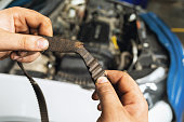 An auto mechanic shows a torn timing belt with worn teeth against the background of an open car hood close-up