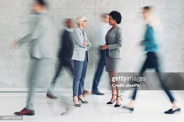 two businesswomen talking while other people walk around in blurred motion - meeting candid office suit stock pictures, royalty-free photos & images