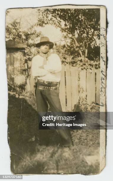 This gelatin silver print depicts a black and white image of a man wearing a hat and gun holster. The man is wearing a long sleeve shirt, pants and a...