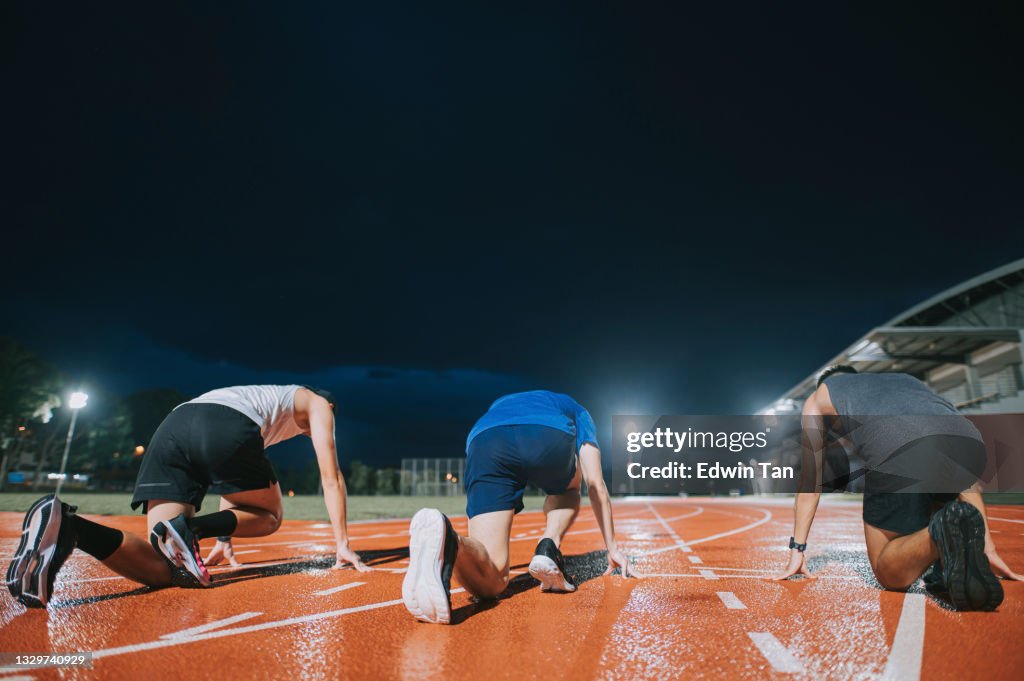 Rear view of asian chinese male athletes lining up starting line aerodynamic shape getting ready to run at track rainy night in stadium