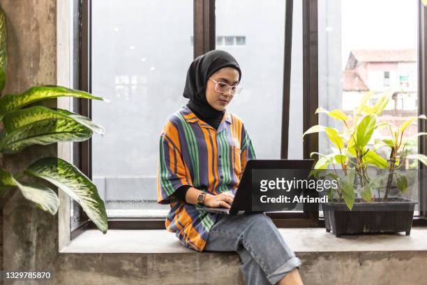 young professional woman working in a co-working space - malay hijab - fotografias e filmes do acervo