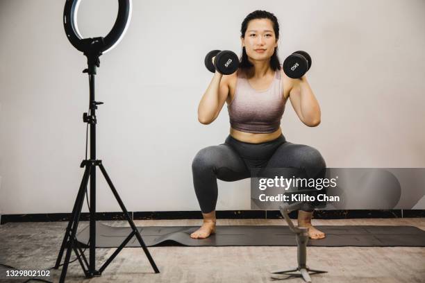 asian woman doing weight training using dumbbell while live streaming via phone on a tripod - asian female bodybuilder stock-fotos und bilder