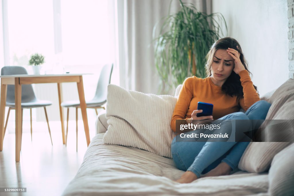 Frustrated and depressed young brunette woman is crying with a smartphone in hands while she sitting on the couch at home
