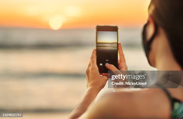 mujer con mascarilla toma una foto de una playa al atardecer - photo messaging fotografías e imágenes de stock