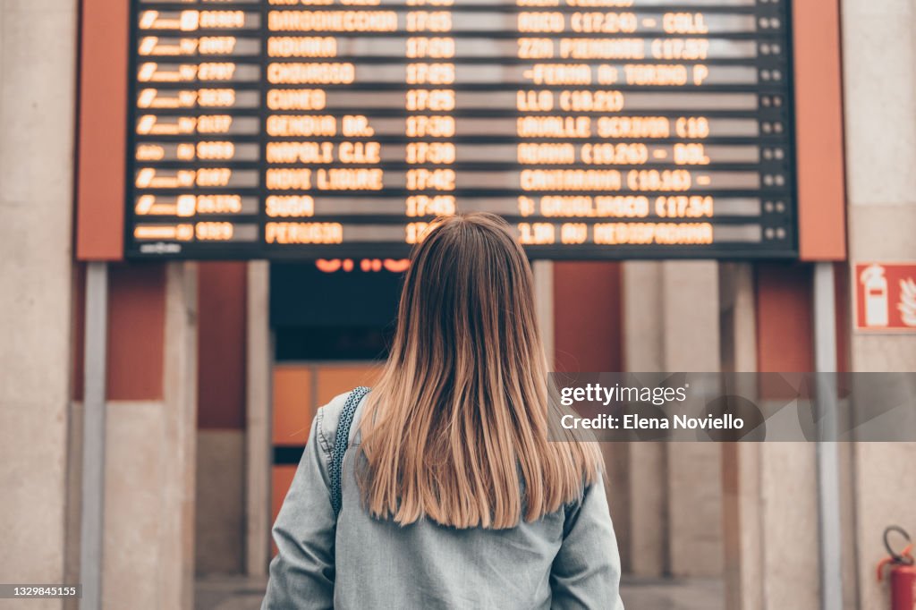 A young woman at a railway station or at the airport looks at the smartphone screen against the background of the arrival and departure board