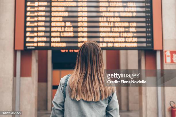 a young woman at a railway station or at the airport looks at the smartphone screen against the background of the arrival and departure board - waiting foto e immagini stock