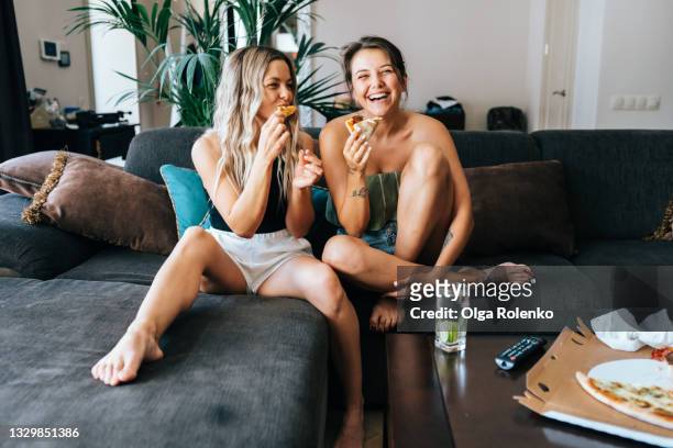 two lesbian women watching tv at home in living room and eating pizza - freunde couch stock-fotos und bilder