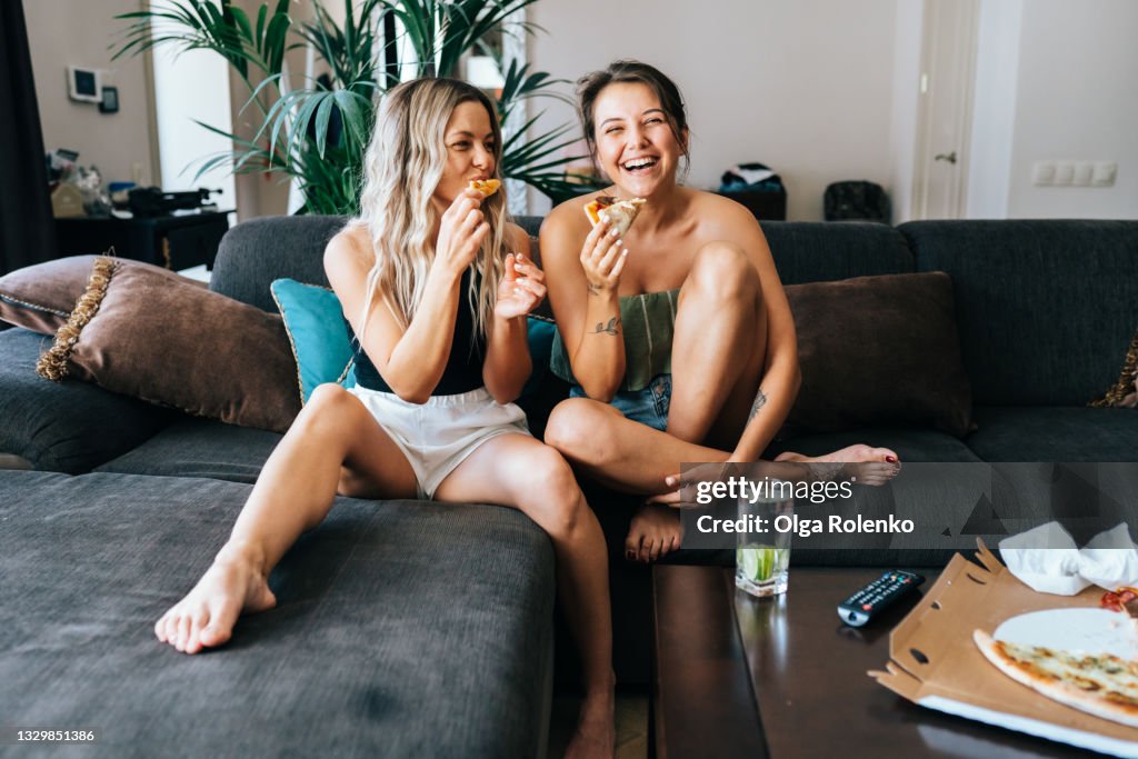 Two lesbian women watching TV at home in living room and eating pizza