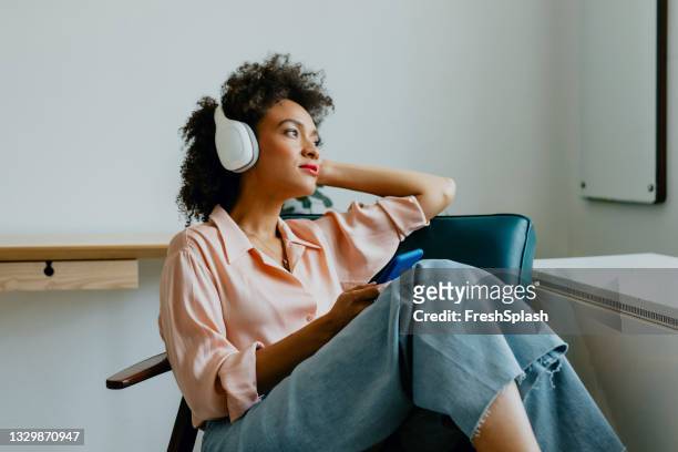 mujer joven relajada con los auriculares puestos, sentada en un sillón y escuchando su podcast favorito - black trousers fotografías e imágenes de stock
