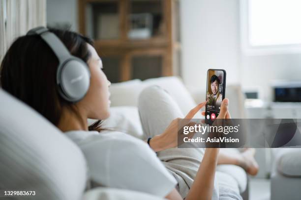 giovane donna asiatica con le cuffie che si rilassano sul divano a casa, utilizzando un'app di incontri online su smartphone, in cerca di amore su internet. social media, incontri su internet, relazione di coppia, concetto d'amore - relazione a distanza foto e immagini stock