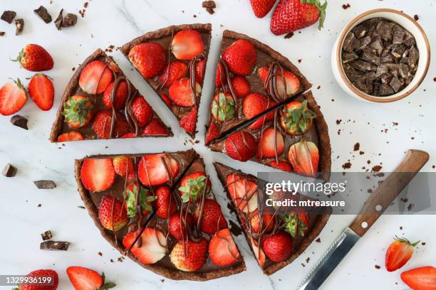 image of sliced, chocolate strawberry tart, crispy, cocoa pastry crust, topped with whole and halved strawberries drizzled with melted chocolate, surrounded by strawberries, beside bowl of chocolate pieces, knife, marble effect background, elevated view - taartpunt stockfoto's en -beelden