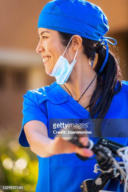 smiling asian mid adult female healthcare worker holding her bicycle outside her home - protective face mask side stock pictures, royalty-free photos & images