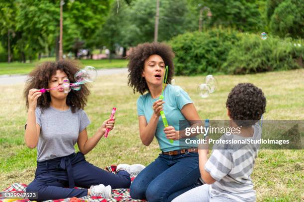 african-american family is sitting on a picnic blanket and blowing bubbles in public park. - preteen model stock pictures, royalty-free photos & images