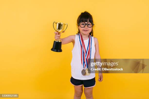 a black-haired girl with glasses, wearing winner medals and lifting a trophy in her hand, on yellow background. olympic and sporting triumph concept. - participant fotografías e imágenes de stock