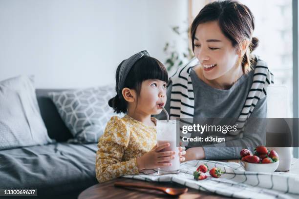 young asian mother making healthy homemade strawberry smoothie with lovely little daughter at home. mother and daughter smiling at each other joyfully and enjoying the drink. healthy eating and healthy lifestyle - milk family stockfoto's en -beelden