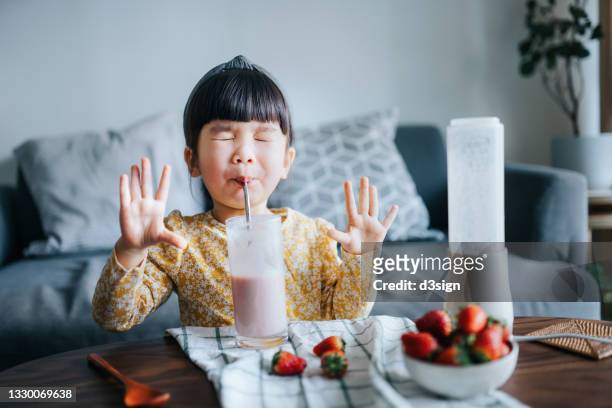 happy little asian girl drinking a glass of healthy homemade strawberry smoothie at home. she is excited with her hands raised and eyes closed while drinking her favourite smoothie. healthy eating and healthy lifestyle concept - healthy refreshment stock pictures, royalty-free photos & images