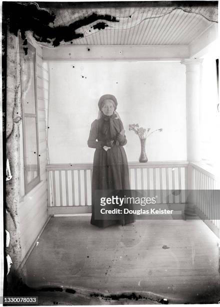 Portrait of an unidentified woman, in hat, gloves, and a long, dark dress as she poses in front of backdrop on a porch, Lincoln, Nebraska, early 20th...